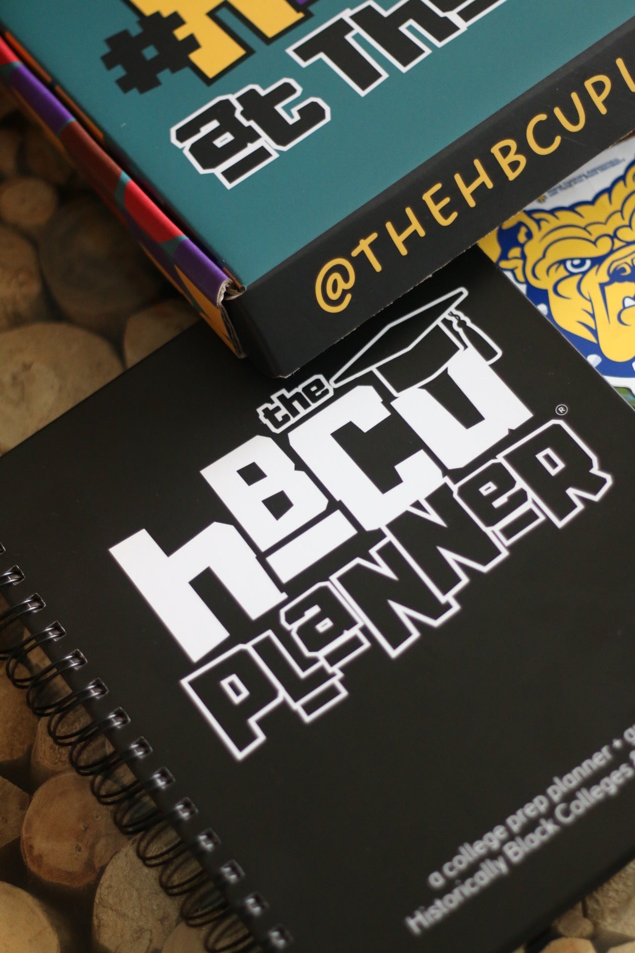 A close-up image of a planner titled 'HBCU Planner' with a black cover and white text. The planner is designed for college planning and is shown with other items, possibly for scale.